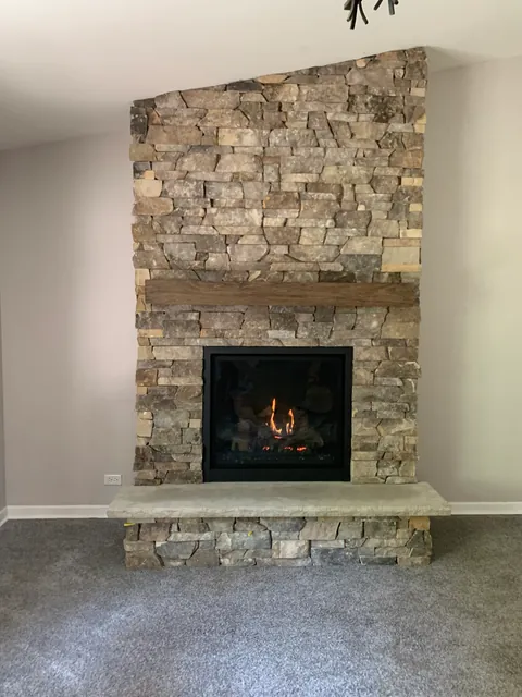 The finished wall: stacked stone from the floor to the sloped ceiling, a wood beam mantel, a raised stone hearth, and the gas fireplace lit.