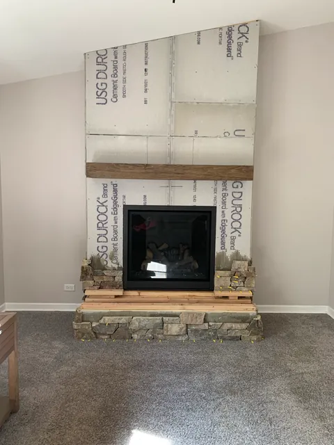 The same wall partway through: cement backer board up to the sloped ceiling, a wood beam mantel in place, and the first rows of stone set around the hearth.