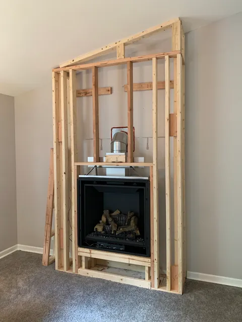 A new gas fireplace set into fresh wood framing against a bare gray wall, with its vent pipe running out through the wall above.