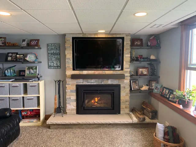 The finished fireplace: a black wood-burning insert with a fire going, set in a stacked stone surround with a mantel shelf and stone hearth.
