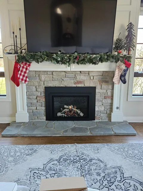 A black gas insert with a birch log set, in a stone surround under a white mantel hung with garland and stockings, a TV mounted above.