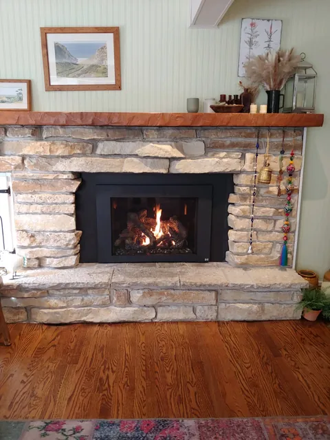 A gas insert with the fire on, set in a light stacked stone fireplace with a raised stone hearth and a rough wood beam mantel.