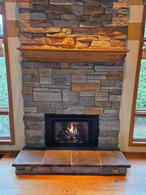 A gas insert with the fire on, in a stone fireplace that runs to the ceiling between two windows, with a wood mantel shelf and a raised stone hearth.