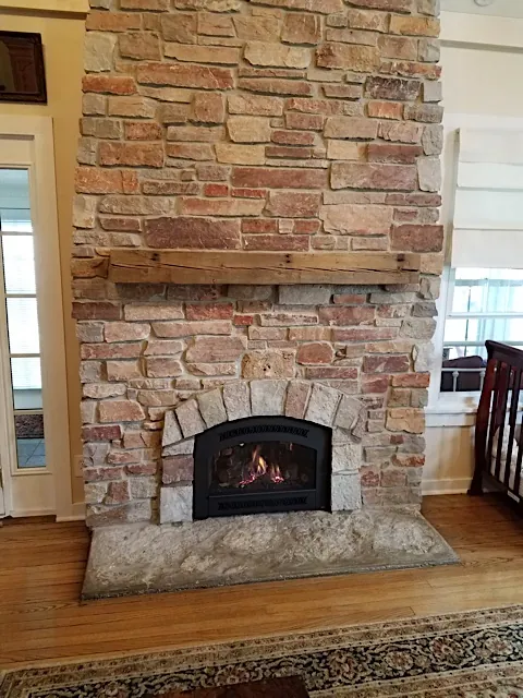A gas insert with an arched front, set under a stone arch in a stone fireplace with a rough beam mantel and a stone hearth.