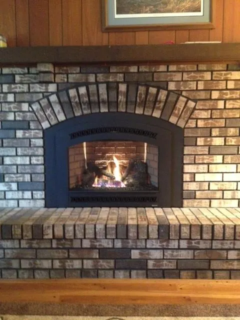 A gas insert with an arched front and the fire on, set under the brick arch of a whitewashed brick fireplace with a raised brick hearth.
