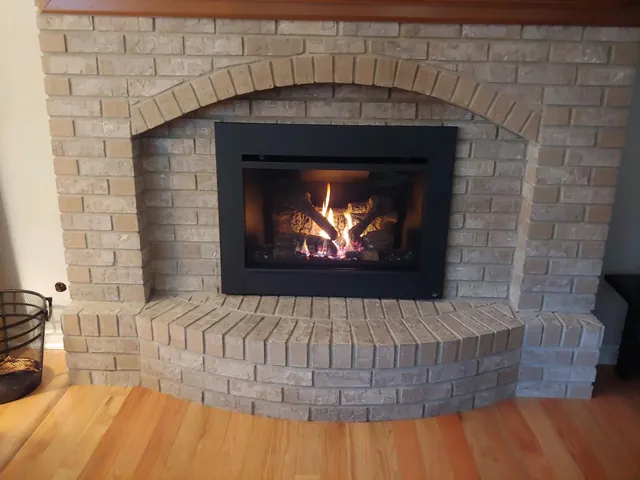 A black gas insert with the fire on, set under the arch of a tan brick fireplace with a curved brick hearth.