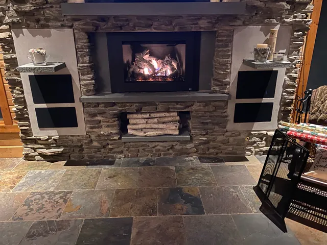 A gas insert with the fire on, set in a stacked stone wall with a shelf on each side and a log nook below, over a slate floor.