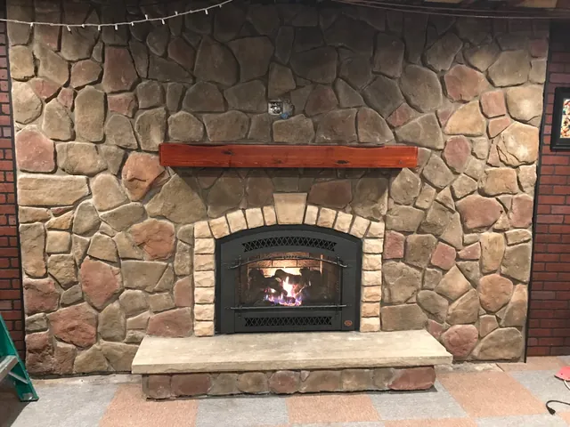 A gas insert with an arched front and the fire on, set in a fieldstone wall under a stone arch, with a red wood mantel and a raised stone hearth.