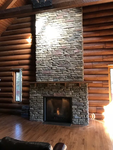 The same log wall with a stacked stone chimney running up to the ceiling beams, a wood mantel, and a glass-front fireplace with a fire lit.