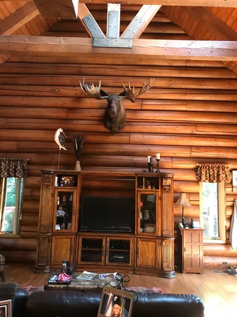 A log home great room with tall log walls, a carved wood entertainment center, and a moose mount on the wall.
